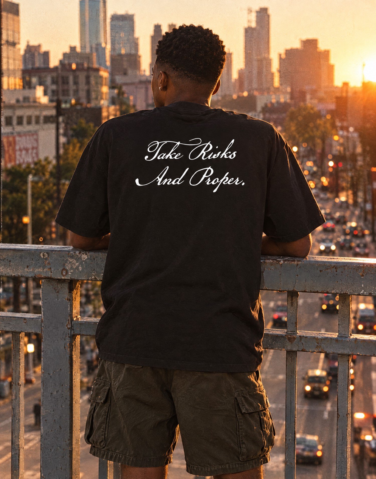 Jeune homme de dos accoudé à une rambarde au-dessus de la ville au coucher du soleil, t-shirt TRAP noir Take Risks And Proper