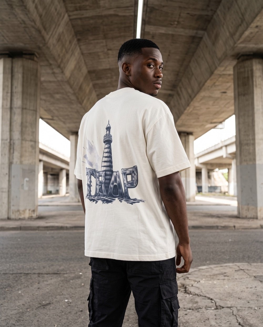 Jeune homme de dos sous un pont, regard par-dessus l'épaule, en t-shirt TRAP blanc avec le phare